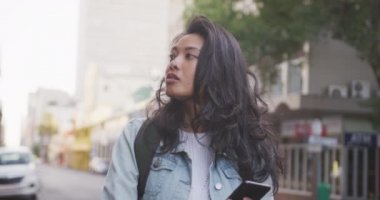 Uzun siyah saçlı, gün boyunca şehir sokaklarında gezen ve arka planda binaları yavaş çekimde olan akıllı telefon kullanan melez bir kadının ön görüntüsü..