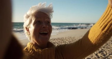 Sahilde güneşin altında kıdemli beyaz bir kadının ön görüntüsü. Kameraya bakıyor ve gülümsüyor. Mavi gökyüzü ve deniz arka planda ağır çekimde.