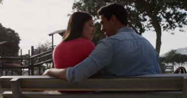 Güneşli bir günde doğada boş zamanların keyfini çıkaran melez bir çiftin yan görüntüsü. Dışarıda bir bankta oturup yavaş çekimde gözleri kapalı oturuyorlar.