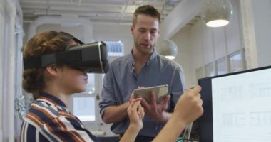  İş adamı ve melez ırk işkadını modern bir ofiste birlikte çalışıyorlar. Sanal gerçeklik kulaklığı takan kadın, arka planda çalışan bayan meslektaşlarıyla birlikte elinde dijital tabletle bir masada oturuyor..