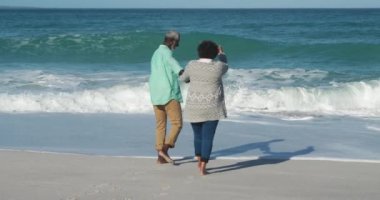 Arka planda mavi gökyüzü ve deniz ile kumsalda vakit geçiren, el ele tutuşan ve yavaşça suya doğru yalınayak yürüyen yaşlı bir Afrikalı Amerikalı çiftin arka plan görüntüsü.