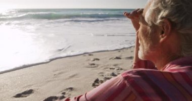 Sahilde kumsalda oturan, yavaşça denize bakan, kıdemli, beyaz bir adamın yan görüntüsü.