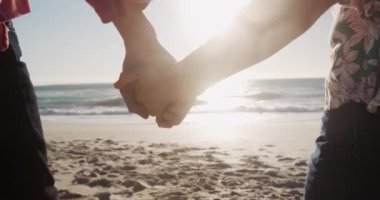 Arka planda mavi gökyüzü ve denizle sahilde yürüyen, ağır çekimde yürüyen ve el ele tutuşan yaşlı beyaz bir çiftin orta kısmı.