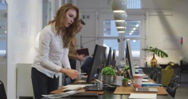 Modern bir ofiste çalışan, masasının başında duran, akıllı telefonuyla konuşan, arka planda iş arkadaşlarıyla bilgisayar başında çalışan beyaz bir iş kadınının yan görüntüsü..