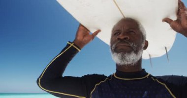 Düşük açılı ön manzara, yaşlı bir Afrikalı Amerikalı 'nın güneşin altında bir sahilde, kafasında mavi gökyüzü ve arka planda sakin bir deniz olan bir sörf tahtası tutarken görüntüsü.