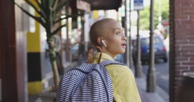 Güneşli bir günde şehirde kısa boyalı sarı saçlı, akıllı telefonuyla yürüyen ve kaykayını taşıyan kulaklıkla müzik dinleyen ve arka planda yavaş çekimde binalara bakan melez bir kadın..