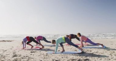 Güneşli bir günde deniz kenarında boş vakit geçiren bir grup beyaz kadın arkadaşın yan görüntüsü. Ağır çekimde yoga alıştırması yapıyorlar.