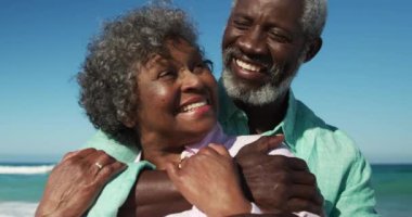 Önde, arka planda mavi gökyüzü ve deniz olan, birbirini kucaklayan ve ağır çekimde gülümseyen mutlu Afrikalı Amerikalı çiftin yakın görüntüsü.