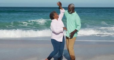 Kıdemli Afrikalı Amerikalı bir çiftin arka planda mavi gökyüzü ve denizle eğlenirken el ele tutuşurken, dans ederken ve ağır çekimde eğlenirken yan görüntüsü.