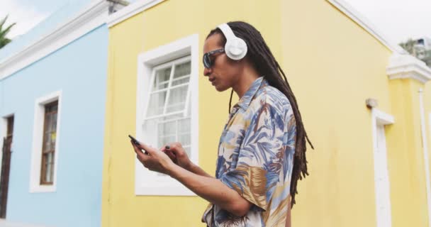 在一个阳光灿烂的日子里 一个长着长发 头戴耳机 戴着太阳镜 站在街上 用智能手机慢动作的混血种族男子的侧影 — 图库视频影像