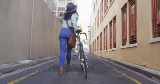 日中は街中や暗い髪の長い混合レースの女性のリアビュー スローモーションの背景に建物と通りに彼女の自転車で歩く — ストック動画