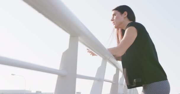 一个健康的白种人女人身穿运动服 穿着黑色长发 在城市的室外锻炼 站在桥上休息 慢动作地拿着一个塑料瓶 侧视图 — 图库视频影像