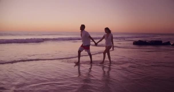 若い混合レースのカップルのサイドビュー日没時に海沿いのビーチで自由な時間を楽しむ — ストック動画