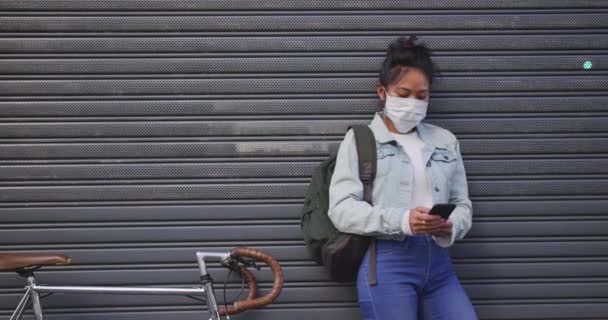 大気汚染やコロナウイルスに対するフェイスマスクを着用し自転車の横に立ちスローモーションを背景に灰色のシャッターを備えたスマートフォンを使用しています — ストック動画