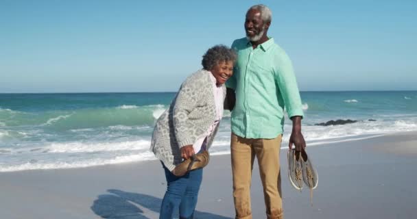 青い空と海を背景にビーチでの時間を楽しんでいるアフリカ系アメリカ人の高齢者のカップルのフロントビュー 裸足で歩くとスローモーションで靴を運ぶ — ストック動画