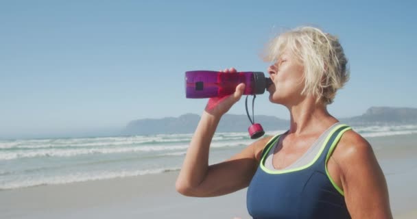 晴れた日に海沿いのビーチで自由な時間を楽しむ先輩白人女性のサイドビュー 運動から休憩を取ります スローモーションでプラスチックボトルから水を飲みます — ストック動画