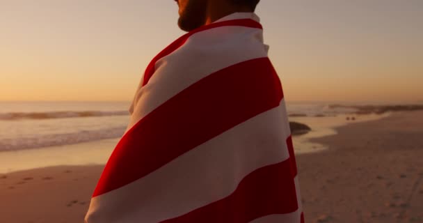 一个年轻的男女混血男子在日落时分在海滩上享受着用美国国旗包裹的闲暇时光 — 图库视频影像