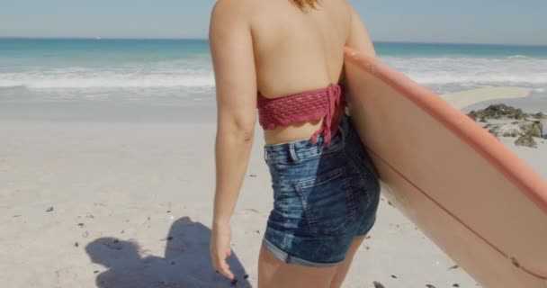 海辺の日当たりの良いビーチでサーフボードと一緒に自由な時間を楽しんでいる若い白人女性の後部ビュー — ストック動画
