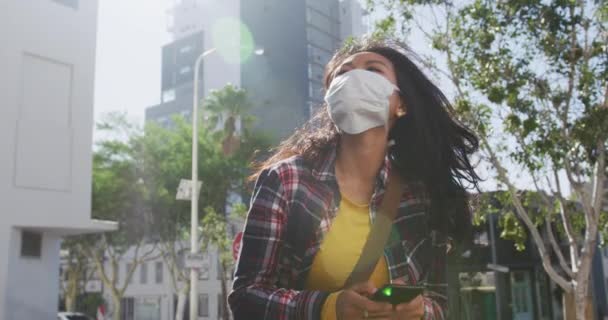 大気汚染やコロナウイルスに対するフェイスマスクを着用し街中を歩きゆっくりとした動きの中で建物を背景にスマートフォンを使い — ストック動画