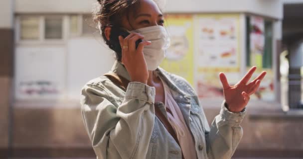 混血的女人 一头乌黑的长发 白天在城市街道上走来走去 戴着防空气污染的面罩 头戴头罩 站在街上 使用智能手机 背景是慢动作的建筑物 — 图库视频影像