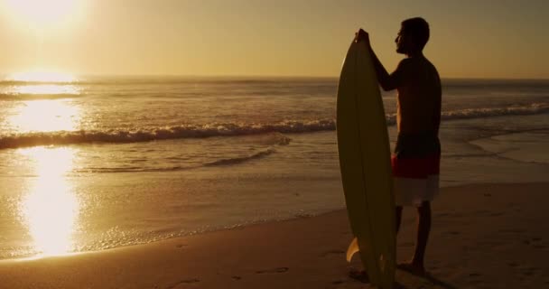 一个年轻的混血种族男子在海滩边享受着与冲浪板在一起和观看日落的闲暇时光 — 图库视频影像