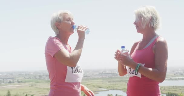 在阳光明媚的日子里 两个白人老年女性朋友一起在乡村享受着闲暇时光 从运动中休息一下 慢吞吞地喝着塑料瓶里的水 在阳光下跑得很开心 — 图库视频影像