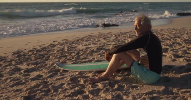 彼のサーフボードの横にある砂の上に座ってゆっくりとした動きで海に目を向ける — ストック動画
