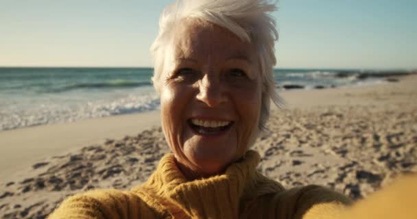 在阳光下的海滩上 一个年长的高加索女人的画像 她看着镜头 微笑着 吹着一个吻 背景是蓝天和大海 动作缓慢 — 图库视频影像