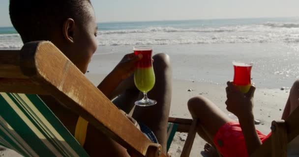 一个年轻的男女混血的男子和一个年轻的非洲裔美国女人坐在海边阳光明媚的海滩上 享受着免费的鸡尾酒和躺椅的侧景 — 图库视频影像
