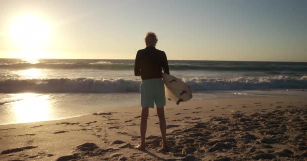 夕阳西下时 一位白人老人站在沙滩上 站在沙滩上欣赏风景 慢吞吞地抱着冲浪板的后视镜 — 图库视频影像