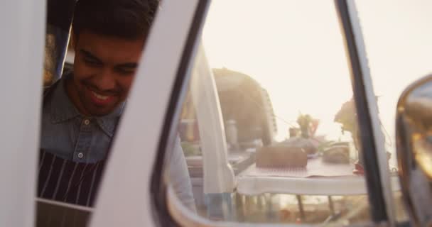 一个混血的男子坐在一辆餐车前面 面带微笑地坐在前面 一个女人从一辆敞着的顶篷面包车后面慢吞吞地吃着东西 — 图库视频影像