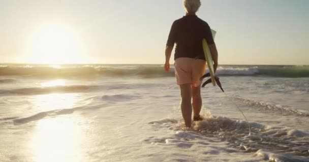 太陽の下でビーチでシニア白人女性の背面ビュー 水の中を歩いて 彼女の腕の下にサーフボードを運ぶ 遅い動きの背景に青い空と海と — ストック動画