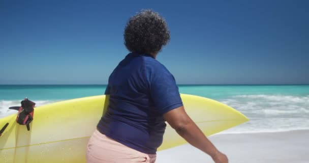 一位非洲裔美国老年妇女在阳光下的海滩上散步 腋下夹着冲浪板 背景为蓝天和大海 动作缓慢的侧视图 — 图库视频影像