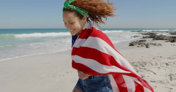 白人の若い女性が海のそばの日当たりの良いビーチでアメリカの国旗と一緒に走る自由時間を楽しんでいる様子 — ストック動画