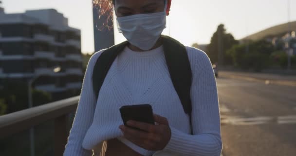 大気汚染やコロナウイルス対策のフェイスマスクを着用し 歩行やスローモーションの背景に都市道路交通を持つスマートフォンを使用して 日中の街の通りに長い暗い髪と約混合レースの女性のフロントビュー — ストック動画