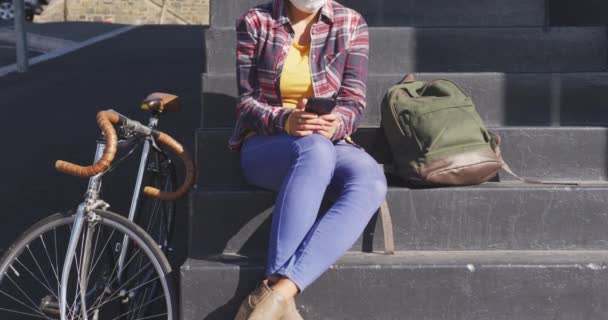 她身披太阳镜 戴着防空气污染面罩 头戴头罩 坐在台阶上 手持智能手机 骑自行车 旁边是交通拥挤的人群 — 图库视频影像