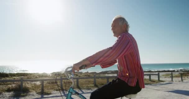 一个白人老人坐在沙滩上晒太阳 骑自行车向前看 背景是蓝天和大海 动作缓慢 — 图库视频影像