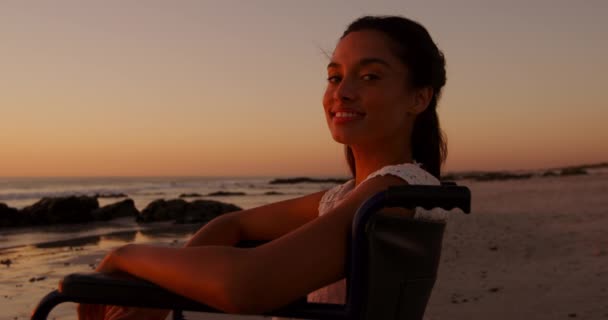 夕暮れ時 海辺のビーチでカメラに笑顔で自由時間を楽しむ車椅子の若い混合レースの女性の肖像 — ストック動画
