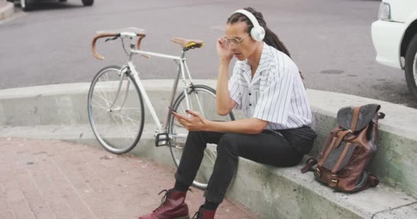 在一个阳光明媚的日子里 坐在街上 头戴耳机 用智能手机 他的自行车慢吞吞地站在他旁边 一个长满了可怕长发的混血赛车手的侧视图 — 图库视频影像