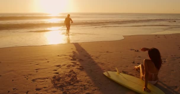 海沿いのビーチで夕日を眺めながら自由な時間を楽しむ若い混合レースカップルのフロントビュー — ストック動画