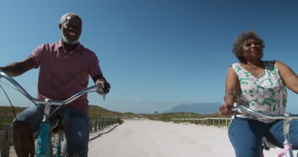 アフリカ系アメリカ人のカップルのサイクリングのフロントビューと遅い動きの背景に青空とビーチで笑顔 — ストック動画