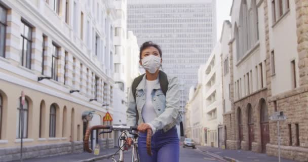 大気汚染やコロナウイルスに対してフェイスマスクを着用し スローモーションの中で建物と彼女の自転車で歩いて 日中の街の通りに長い暗い髪と約混合レースの女性のフロントビュー — ストック動画