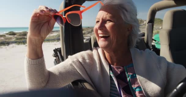 道路の旅の間に太陽の下でビーチでシニア白人女性の側のビューは 車の運転席に車の後ろに座って サングラスを着用し サイドウィンドウのスローモーションで笑みを浮かべて外を見る — ストック動画
