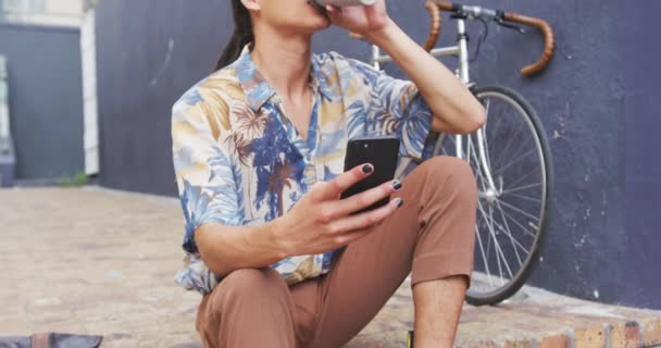 道路に座ってコーヒーを飲んでスマートフォンを使って自転車でゆっくりと動きながら横の壁にもたれています彼は車を運転しました — ストック動画