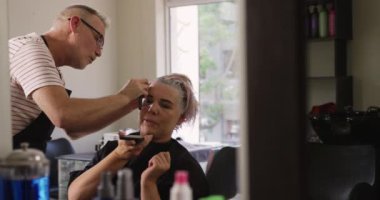 Alternatif havalı kuaför salonu. Kafkasyalı bir erkek kuaförün kuaförde çalıştığını, elinde jilet tuttuğunu, beyaz bir kadının saçını tıraş ettiğini, akıllı telefon kullandığını, aynada, yavaş çekimde yansıttığını.