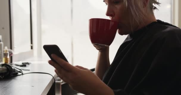 代替クールなヘアサロン ヘアサロンに座っている白人女性の顧客のサイドビュー コーヒーを飲みます スマートフォンを使用して スローモーションで — ストック動画