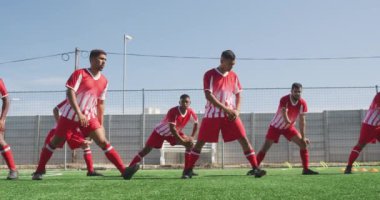 Çok ırklı bir grup erkek futbolcunun takım elbiseli, güneşin altında bir spor sahasında antrenman yapan, esneyen, ağır çekimde