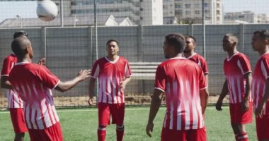 Çok ırklı erkek futbol oyuncularından oluşan bir grubun ön görüntüsü. Takım elbisesi giyiyor, güneşin altında bir spor sahasında antrenman yapıyor, yavaş çekimde topu tekmeliyor ve pas veriyor.
