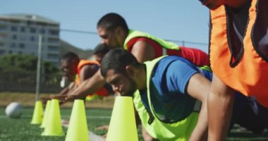 Spor giyim ve renkli önlük giyen, spor sahasında antrenman yapan, peş peşe şınav çeken ve ağır çekimde konilere dokunan çok ırklı erkek futbol oyuncularının ön görüntüsü.