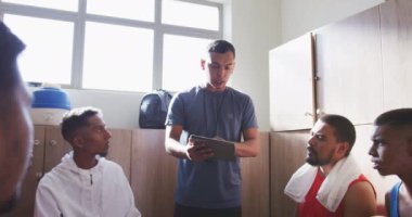 Spor kıyafetleri giyen, soyunma odasında oturan, karışık yarış koçlarının elinde bir pano tutan ve yavaş çekimde motive eden çok ırklı erkek futbol oyuncularının ön görüntüsü.